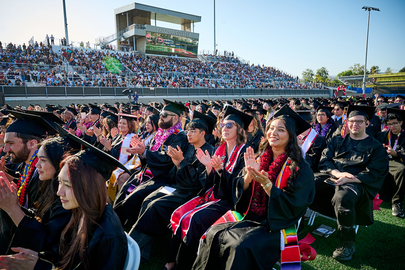 Graduates seated at an outdoor ceremony with a large crowd.